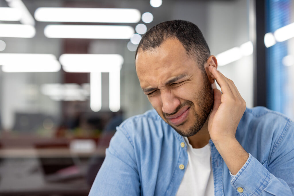 Close-up photo of a young Latin American man in the office, grimacing in pain, holding his ear with his hand.